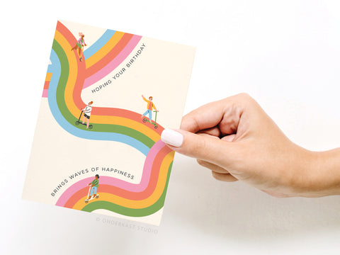 A hand holds the Onderkast Studio Waves of Happiness Birthday Greeting Card, featuring vibrant wavy rainbow stripes, people on scooters and bikes, and the message: Hoping your birthday brings waves of happiness.