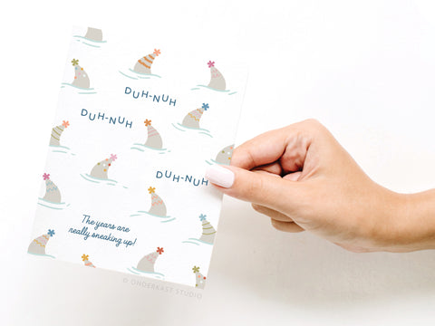 A hand with light pink nail polish holds Onderkast Studio’s Shark Fin Party Hats Birthday Greeting Card, featuring illustrated sharks in party hats, with DUH-NUH repeated and “The years are really sneaking up!” at the bottom.