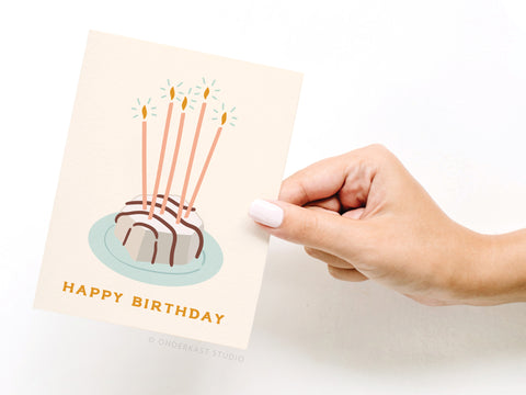 A hand with light pink nail polish holds the Onderkast Studio Happy Birthday Zebra Cake Greeting Card, featuring a whimsical cake with five candles and HAPPY BIRTHDAY in yellow at the bottom.