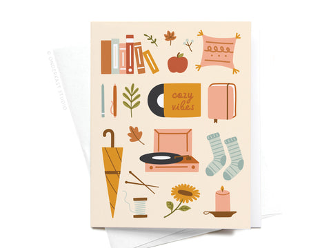 The Cozy Vibes Greeting Card by Onderkast Studio features vibrant illustrations of fall-themed items—books, a pillow, apple, and a mug labeled Cozy Vibes—for a premium touch. Ideal as seasonal decor or to share cozy greetings.
