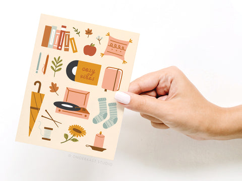 A hand holds the Cozy Vibes Greeting Card by Onderkast Studio, showcasing vibrant autumn-themed prints—books, socks, a candle, tea, and a record player—designed to evoke premium comfort and warmth.