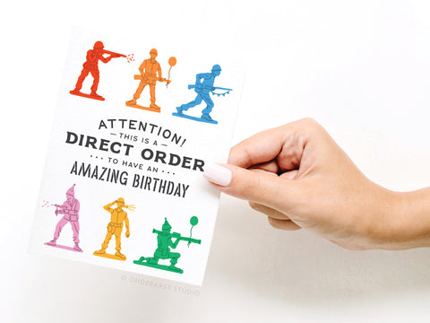 A hand holds the Onderkast Studio Direct Order Army Men Birthday Greeting Card, featuring colorful toy soldier art and the message: “Attention! This is a direct order to have an amazing birthday.”.