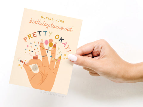 A hand holds the Pretty Okay Birthday Greeting Card by Onderkast Studio, featuring “Hoping your birthday turns out pretty okay!” and an illustration of a hand with a cupcake, balloons, and party confetti.