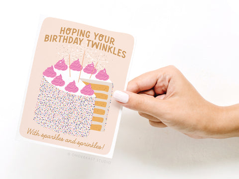 A hand holds the Onderkast Studio Birthday Sparkles and Sprinkles Greeting Card, which features a cake slice with pink frosting, sprinkles, sparklers, and the message: Hoping your birthday twinkles with sparkles and sprinkles!.