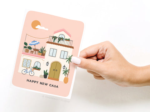 A hand with white-painted nails holds the Onderkast Studio Happy New Casa Greeting Card, featuring an illustrated house with balconies, plants, a bicycle, and “HAPPY NEW CASA” text on a pastel pink background.