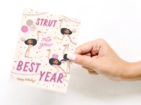 A hand with light pink nail polish holds the Strut Into Your Best Year Ostrich Greeting Card by Onderkast Studio, featuring an ostrich with balloons, confetti, and presents. The card reads: STRUT into your BEST YEAR. happy birthday! in pink and gold letters.