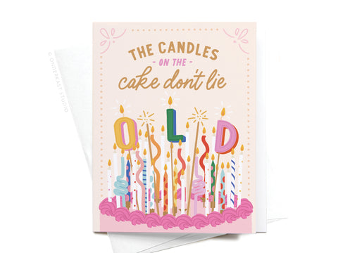 The Candles on the Cake Don’t Lie Greeting Card by Onderkast Studio features candles spelling “OLD” on a pink cake, confetti, stars, and playful text: “THE CANDLES ON THE cake don’t lie.” Includes a partially visible envelope.