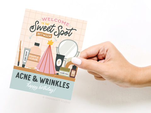 A hand holds the Sweet Spot Between Acne & Wrinkles Greeting Card by Onderkast Studio, featuring skincare products, a mirror, and a pink cosmetic bag. The card says: Welcome to the sweet spot between acne & wrinkles. Happy birthday!.