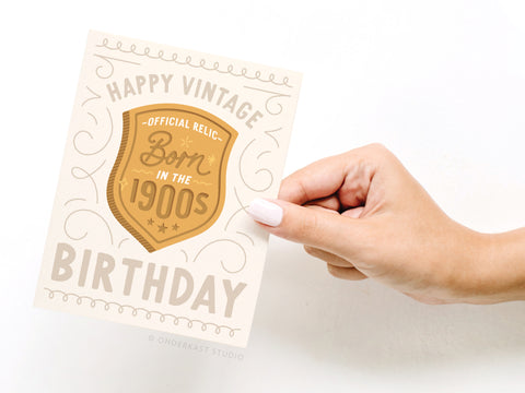 A hand with light nail polish holds the Onderkast Studio Born in the 1900s Vintage Birthday Greeting Card, featuring “HAPPY VINTAGE BIRTHDAY,” “Official Relic: Born in the 1900s,” decorative swirls, and a gold shield.