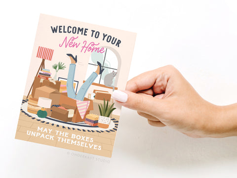 A hand holds the Onderkast Studio Welcome to Your New Home Boxes Greeting Card, featuring legs sticking out of moving boxes and a cat, with the message: “Welcome to your new home. May the boxes unpack themselves.”.