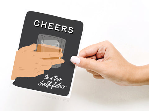 A hand holds Onderkast Studio’s “Cheers to a Top Shelf Father” greeting card, which features an illustration of a hand raising a glass of whiskey and the message CHEERS to a top shelf father on a black background—perfect for Fathers Day.