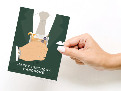 A hand with white-painted nails holds the Happy Birthday Handsome Greeting Card by Onderkast Studio, featuring a man in a suit with a cocktail and the message: HAPPY BIRTHDAY, HANDSOME.