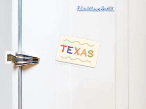 A white retro-handle fridge displays an Onderkast Studio Funky Texas Refrigerator Magnet with a vibrant design, plus a blue Elektrobit magnet, adding Lone Star State flair to the door.