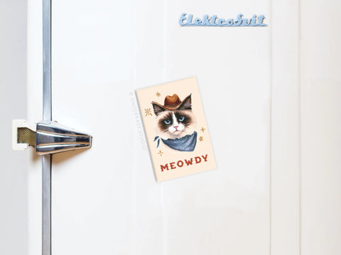 A refrigerator door with a silver handle and a blue Elektrobit magnet at the top. An Onderkast Studio Meowdy Cowboy Cat Refrigerator Magnet shows a grumpy cat in a cowboy hat and bandana labeled “MEOWDY.”.