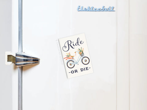 A white fridge with a silver latch features a blue “Elektrobit” magnet and the Ride Or Die Bicycle Refrigerator Magnet from Onderkast Studio, which holds a card reading “Ride -or die-” above a vintage-style bicycle with flower baskets.