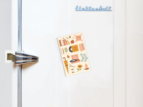 A white fridge with an Elektrobit logo and silver handle displays the Cozy Vibes Refrigerator Magnet by Onderkast Studio, a vibrantly printed decorative card with autumn-themed art like books, socks, leaves, and a pumpkin.