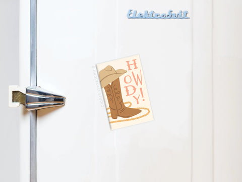 A white refrigerator with a metal handle and the Elektrobit logo displays the Onderkast Studio Howdy Boot Refrigerator Magnet, featuring a cowboy boot, hat, lasso, and “HOWDY!” in bold letters.