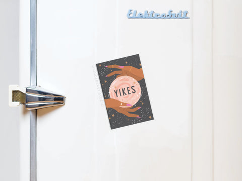 A whimsical Yikes Fortune Teller Refrigerator Magnet by Onderkast Studio, shaped like a crystal ball held by two hands, secures a postcard to a white fridge door with a vintage silver handle.