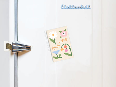 A Bloom & Grow Refrigerator Magnet by Onderkast Studio, featuring a floral design and uplifting message, decorates the white fridge door. A blue Elektrobit logo appears on the top right of the refrigerator.
