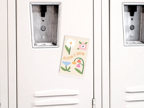 A pastel-toned Bloom & Grow Refrigerator Magnet by Onderkast Studio with a positivity floral design and inspiring words decorates a light gray locker with ventilation slots and a lock mechanism.