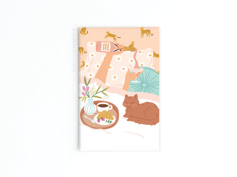 The Rise & Wine Refrigerator Magnet by Onderkast Studio features a cozy bed with patterned pillows, a teal cushion, a sleeping brown cat, and a breakfast tray with coffee, croissant, wine, and flowers on a soft peach cheetah-print background.