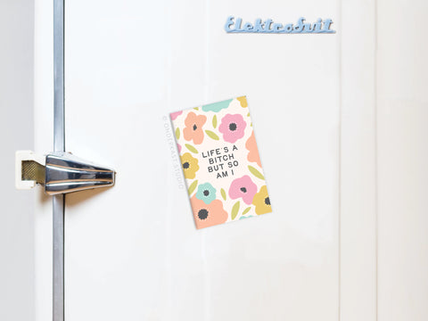 The Life’s a Bitch But So Am I Refrigerator Magnet by Onderkast Studio, featuring a pastel floral design and empowering message, is displayed on a white fridge door with a silver handle to the left. A blue Elektrobit magnet sits above it.