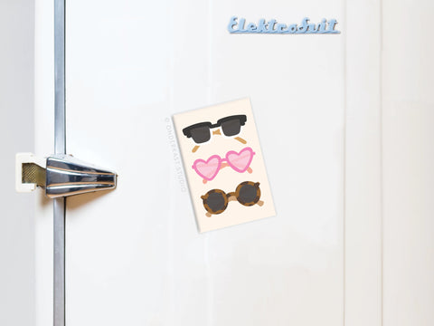 A postcard of three sunglasses—black, pink heart-shaped, and tortoiseshell—pairs with the blue Onderkast Studio Sunglasses Refrigerator Magnet reading Elektrobit for a vibrant display on a white fridge.