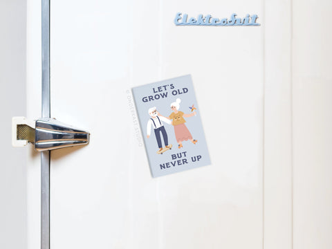 The Onderkast Studio “Let’s Grow Old But Never Up Refrigerator Magnet” features cartoon elderly couples holding hands and a card that reads, LETS GROW OLD BUT NEVER UP.