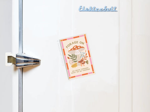 A vintage white fridge with a metal handle displays a blue Elektrobit magnet and the Forage On Mushrooms Refrigerator Magnet by Onderkast Studio, featuring mushrooms, leaves, and the quote: FORAGE ON - WE GROW THROUGH WHAT WE GO THROUGH.