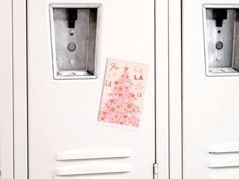 A Fa La La Pink Christmas Tree Refrigerator Magnet by Onderkast Studio is taped to a white locker door between two metal handles, resembling a pink Christmas card with festive text and a decorated tree.