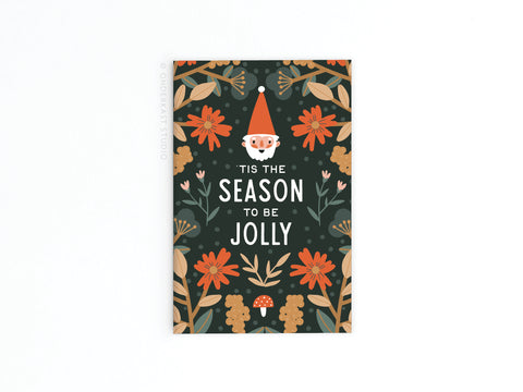The Onderkast Studio Tis the Season To Be Jolly Refrigerator Magnet features a gnomes face and festive decorations—orange flowers, leaves, and a red mushroom—on a dark background, perfect for spreading holiday cheer.