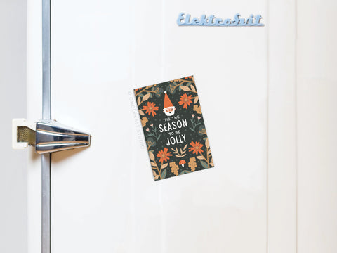 The ’Tis the Season To Be Jolly Refrigerator Magnet by Onderkast Studio displays a festive floral design and cheerful holiday message as it holds a card on a white refrigerator door.