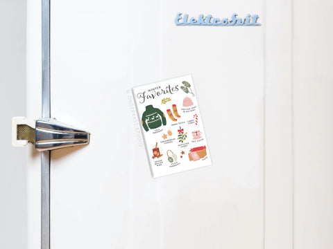 A white fridge with a silver handle displays the Onderkast Studio Winter Favorites Refrigerator Magnet, featuring cozy items like a sweater, tea, and socks—a charming holiday decor piece.
