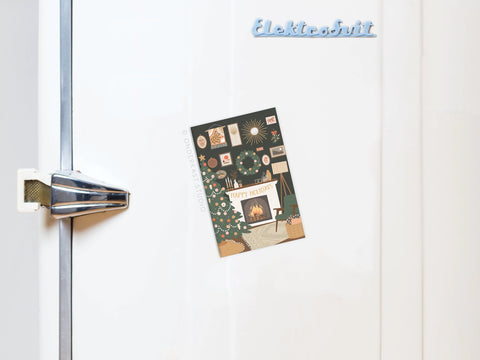 The Happy Holidays Cozy Fireplace Refrigerator Magnet by Onderkast Studio features a festive Christmas tree and fireplace design, attached to a white fridge with Elektrobit on top. The fridge handle is visible on the left.
