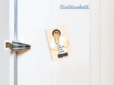 An Onderkast Studio TS Red Refrigerator Magnet holds a postcard of an illustrated woman in sunglasses and a hat with the words Not a lot going on at the moment, placed near a metallic handle and Elektrobit logo.