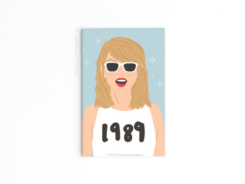 Show off your love for pop perfection with Onderkast Studio’s TS 1989 Refrigerator Magnet, featuring a blonde woman in white sunglasses and a “1989” top on a starry light blue background.