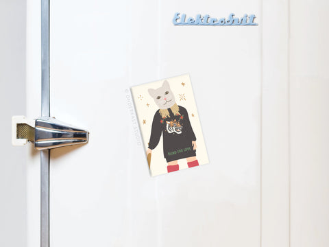 A postcard of a cat in a sweater is attached to a white fridge with a metal handle. An Onderkast Studio TS Reputation Refrigerator Magnet inspired by Taylor Swift adds charm, as “Elektrobrät” appears in blue at the top right.