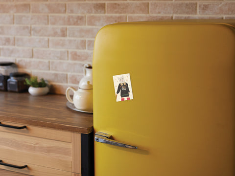 A yellow retro-style refrigerator in a cozy kitchen is adorned with the TS Reputation Refrigerator Magnet by Onderkast Studio, inspired by Taylor Swift’s LWYMMD era. The scene includes a wooden countertop, teapot, cups, and brick-patterned wall tiles.