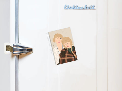 A white fridge with an Onderkast Studio logo and metallic handle, featuring the TS Folkevermore Refrigerator Magnet—an illustration of two blondes (one in a checkered coat) leaning together, inspired by Taylor Swift’s folklore and evermore eras.