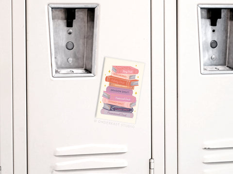 The Smut Book Stack Refrigerator Magnet by Onderkast Studio features pastel-hued books with gold accents—a playful design perfect for any bookworms locker or fridge.