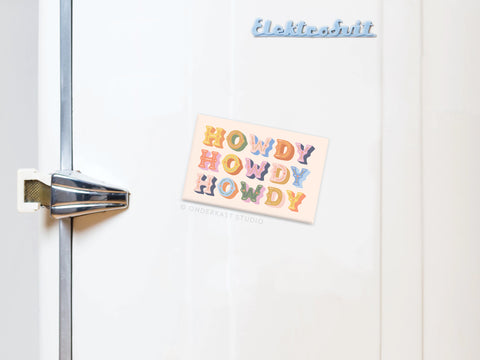 A white refrigerator with a silver handle features an Onderkast Studio Howdy Howdy Howdy Refrigerator Magnet, printed in the USA, and a blue Elektrosvit refrigerator magnet.