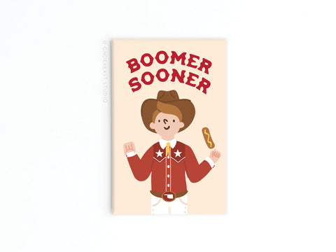 The Boomer Sooner Refrigerator Magnet by Onderkast Studio features a smiling cowboy in a red shirt and brown hat holding a corndog, with bold Boomer Sooner lettering—fun OU gear for Oklahoma fans or as a Big Tex-inspired fridge magnet.