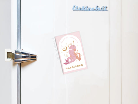 The Capricorn Zodiac Refrigerator Magnet by Onderkast Studio holds a postcard with a pink goat and celestial charm on a white fridge, featuring “Elektrosvit” in blue at the top and a metal latch on the door.