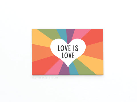The Love Is Love Refrigerator Magnet by Onderkast Studio is heart-shaped and features LOVE IS LOVE over a rainbow burst with vibrant red, orange, yellow, green, blue, and pink rays—symbolizing equality and inclusion.