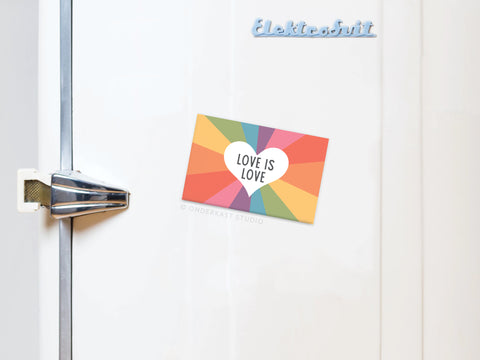 The Love Is Love Refrigerator Magnet by Onderkast Studio features a heart with rainbow rays and the message LOVE IS LOVE, celebrating equality and inclusion. It’s shown on a white fridge with vibrant colors radiating from the design.