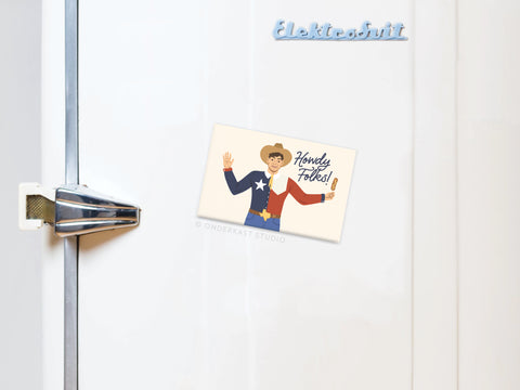 A white fridge with a chrome latch handle, topped with light blue “Elektrobit” letters. The Onderkast Studio Big Tex Refrigerator Magnet depicts a smiling cowboy waving and “Howdy Folks!” in bold, vibrant colors.