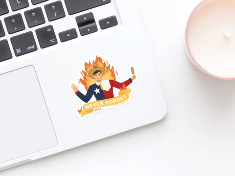 A laptop sports the Onderkast Studio Never Forget Blazing Big Tex Sticker—showing a corn dog-wielding figure in a Texas flag shirt, arms raised amid flames, with “NEVER FORGET 2012” capturing this vivid moment in Texas history. A candle sits nearby.