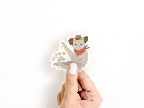 A hand holds the Meowdy Sticker by Onderkast Studio—a die cut vinyl of a gray cowboy cat in a hat and red bandana, with MEOWDY by its tail. Perfect for cowboy cat fans, it pops against any white background.