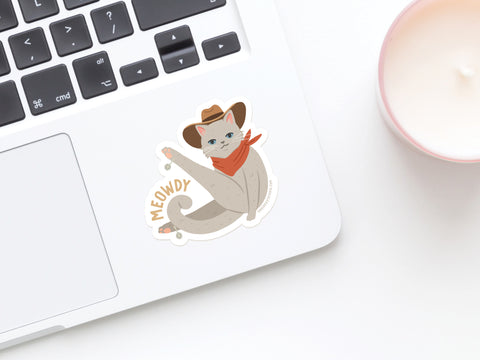 The Meowdy Sticker from Onderkast Studio shows a gray cowboy cat in a hat and red bandana with MEOWDY text, adding quirky charm to a white laptop next to a pink-rimmed candle. Ideal for fans of unique cowboy cat designs!.