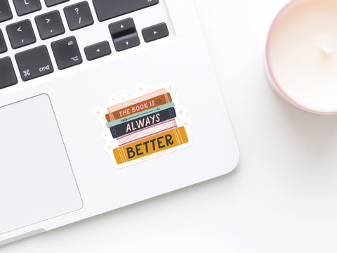 A laptop with The Book Is Always Better Sticker by Onderkast Studio—a vinyl sticker of a book stack—beside a pink-edged candle on a white surface. Printed in the USA.